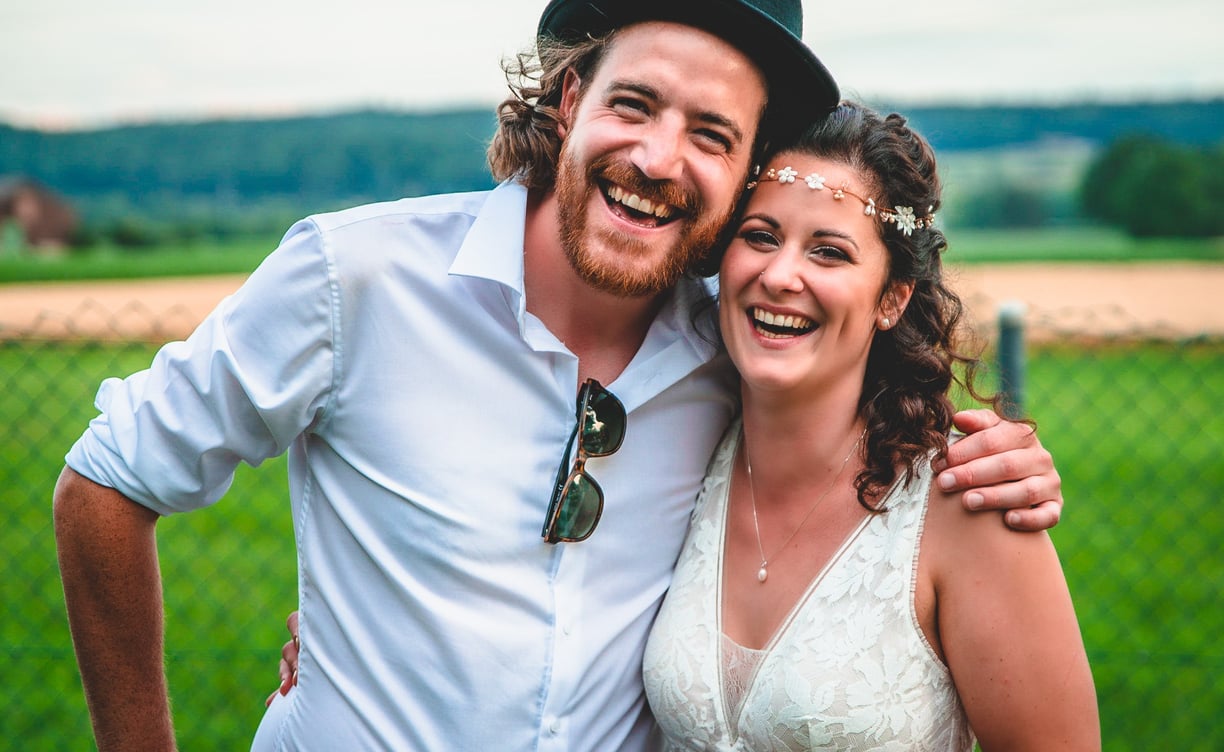Bräutigam mit Hut und Braut mit Haarband lachen vor der Landschaft – Hochzeit von Rege & Sven