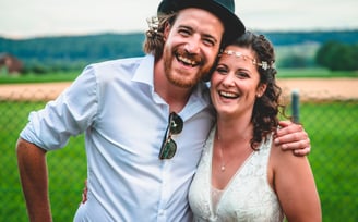 Bräutigam mit Hut und Braut mit Haarband lachen vor der Landschaft – Hochzeit von Rege & Sven