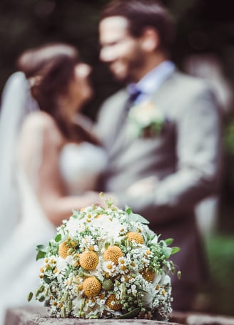 Brautstrauss mit gelben Blüten, Brautpaar im Hintergrund – Hochzeit von Misch & Simu
