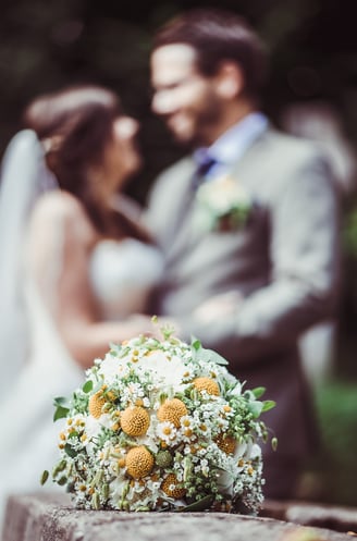 Brautstrauss mit gelben Blüten, Brautpaar im Hintergrund – Hochzeit von Misch & Simu