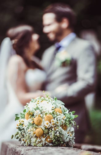 Brautstrauss mit gelben Blüten, Brautpaar im Hintergrund – Hochzeit von Misch & Simu
