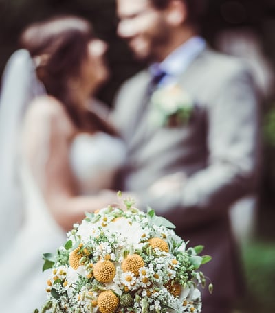 Brautstrauss mit gelben Blüten, Brautpaar im Hintergrund – Hochzeit von Misch & Simu
