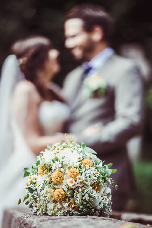 Brautstrauss mit gelben Blüten, Brautpaar im Hintergrund – Hochzeit von Misch & Simu