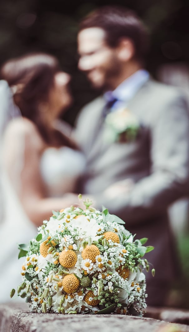 Brautstrauss mit gelben Blüten, Brautpaar im Hintergrund – Hochzeit von Misch & Simu